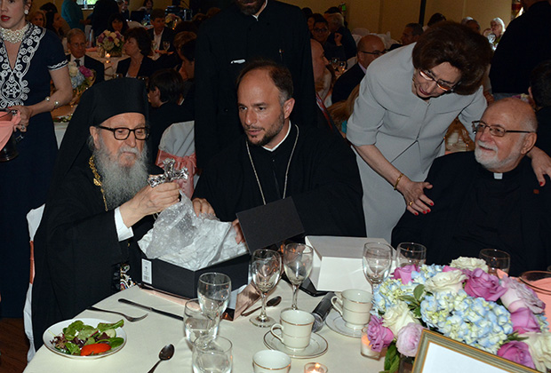 Archbishop Demetrios is unwrapping a thank you gift offered by the community, while Deacon Panteleimon, Fr. Elias Villis, Fr. Nicholas Triantafilou, until recently President of the Hellenic College/Holy Cross School, and Paulette Poulos, Executive Director of Leadership 100, are watching